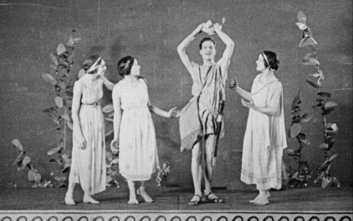 Black-and-white archival image depicting four actors dressed in Greco-Roman-inspired robes within a theatrical stage set.