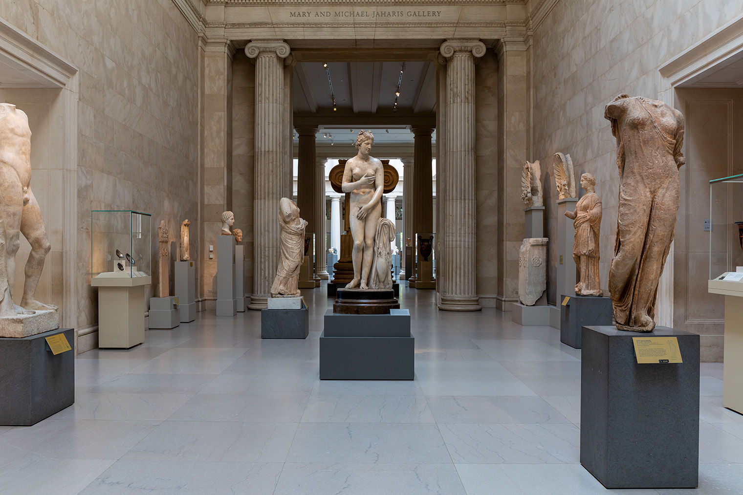 A marble statue of Aphrodite stands in the middle of the Greek and Roman hall surrounding by other statues and sculptures. She is standing upright with her hands moving to cover her breasts and pubic area. 