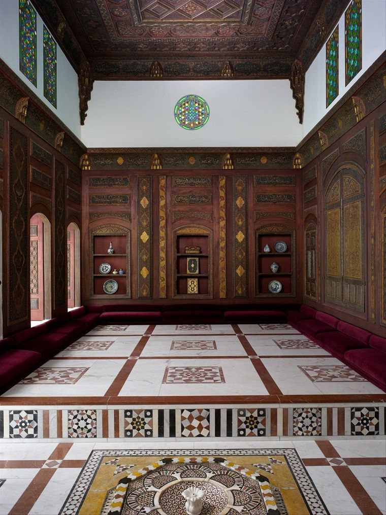 A room from Damascus decorated with gilded dark wood paneling, low dark red velvet covered cushions; open windows on the left illuminate the room with light
