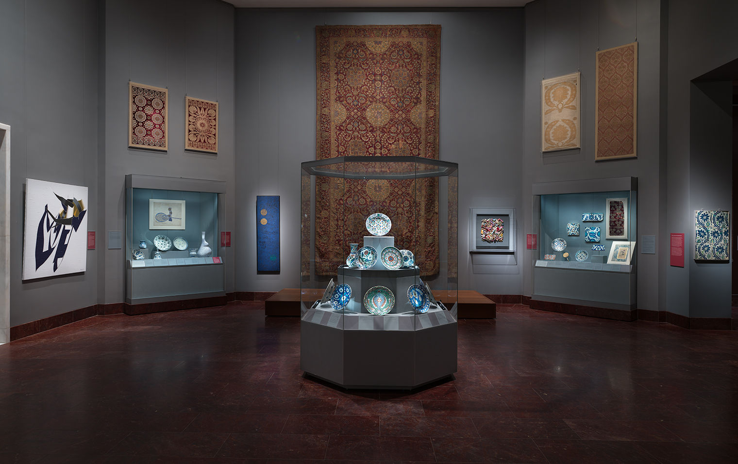 Gallery view of the installation, Dialogues: Modern Artists and the Ottoman Past, in the Koç Family Galleries - Carpets, Textiles and the Greater Ottoman World and Arts of the Ottoman Court (14th–20th centuries) depicting a central glass case with Ottoman ceramics in the foreground, and wall cases, carpets and textiles, and modern artworks on the walls in the background