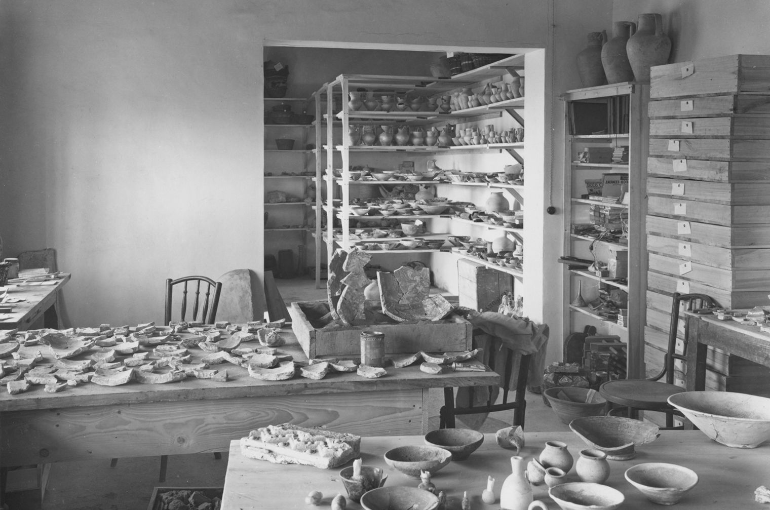 Black and white photo with tables in the foreground and wall shelves in the background with scattered ceramics and ceramic fragments