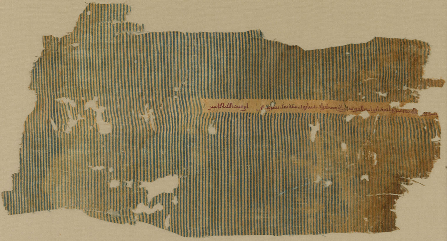 A silk and cotton tiraz textile fragment with thin vertical stripes and a thin line of embroidered red calligraphy on the right side of the textile