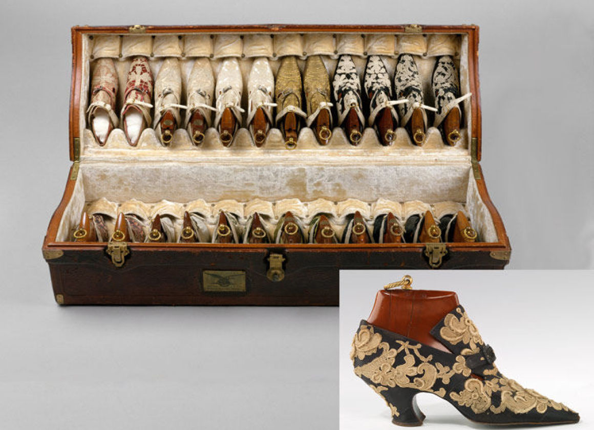 Composite image: in the foreground, a low-heeled, pointed shoe with a wooden insert appears in profile against a seamless off-white background; in the background, an open suitcase containing pairs of shoes that are also filled with wooden inserts rests agains a greyish seamless background
