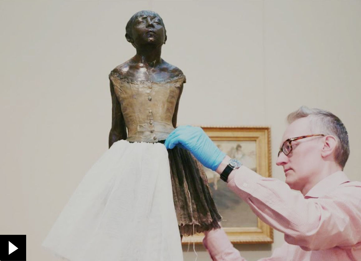 A conservator wraps a skirt around a sculpture of a ballerina dancer by Degas in the Impressionist painting galleries; there is a small play button icon in the bottom left-hand corner