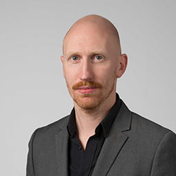 Light skinned middle-aged man with shaved head, ginger moustache and stubble beard wearing a black shirt and grey suit jacket 