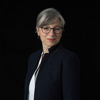 Smiling light-skinned woman with grey hair and glasses wearing a white shirt and a dark-blue blazer 