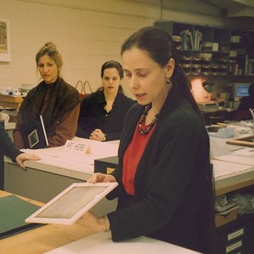 A conservator holds what appears to be a white framed or matted print over a large island table towards a group of observers. Two spectators look on from behind.