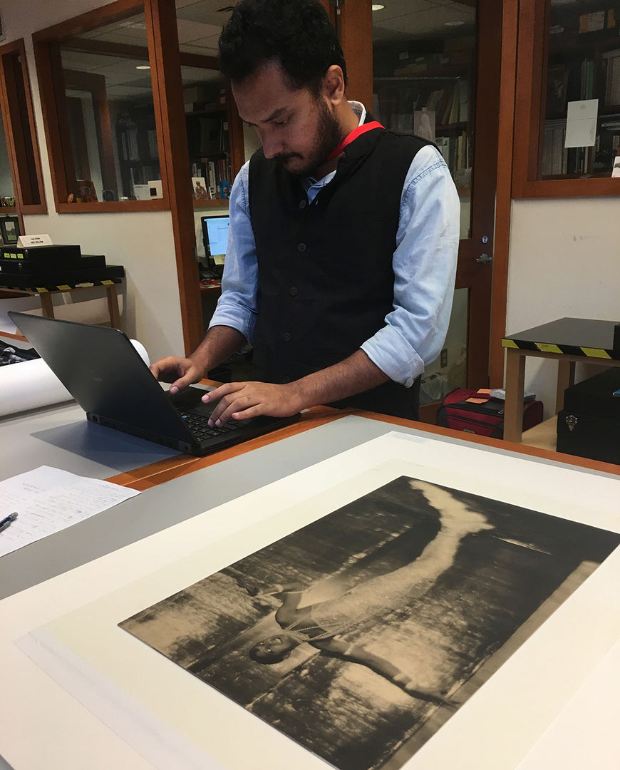 A Met Museum Fellow stands at a work table typing on laptop, with a photograph laying on the tabletop in the foreground.