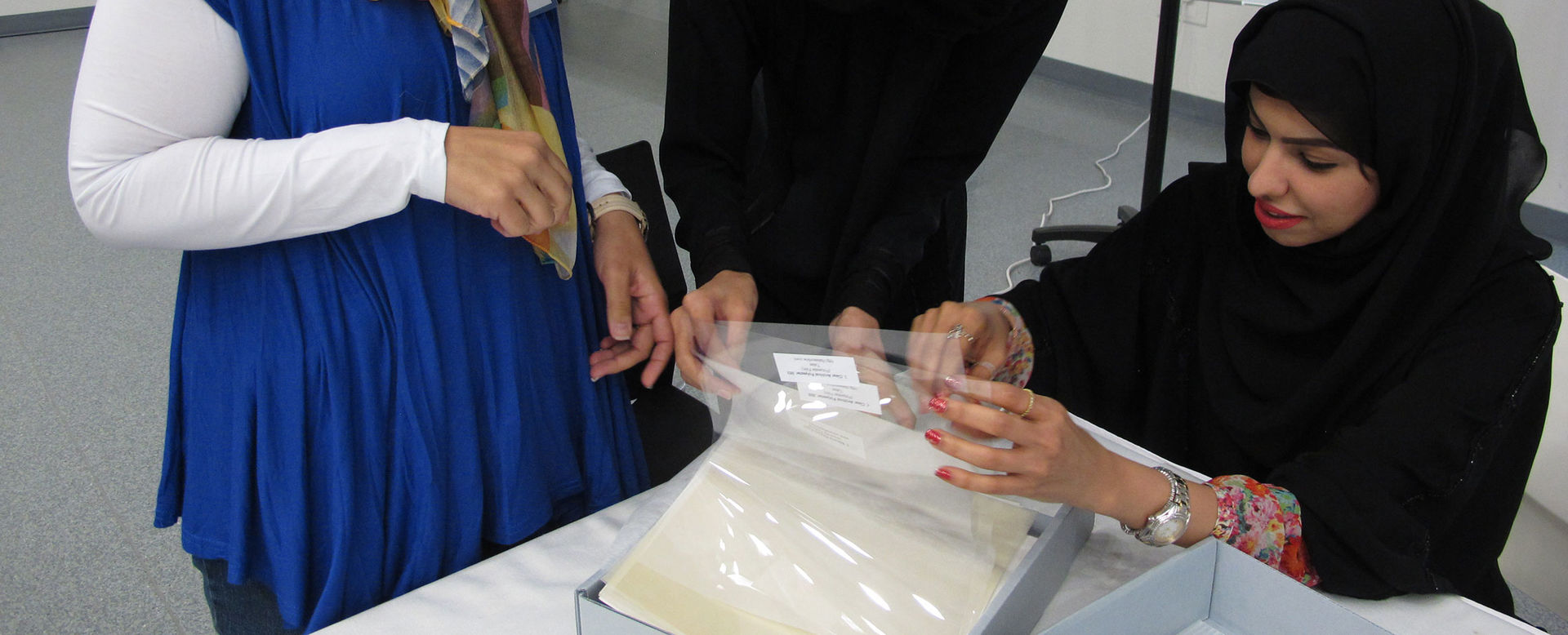 A group of women gather round a table to leaf through and consult papers protected in plastic sleeves stored in archival boxes