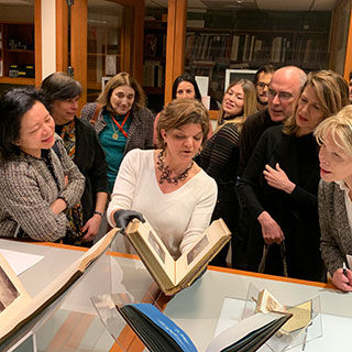 The conservator, Georgia Southworth delicately holds a large album of prints or illustrations slightly ajar to a group of spectators surrounded behind her.