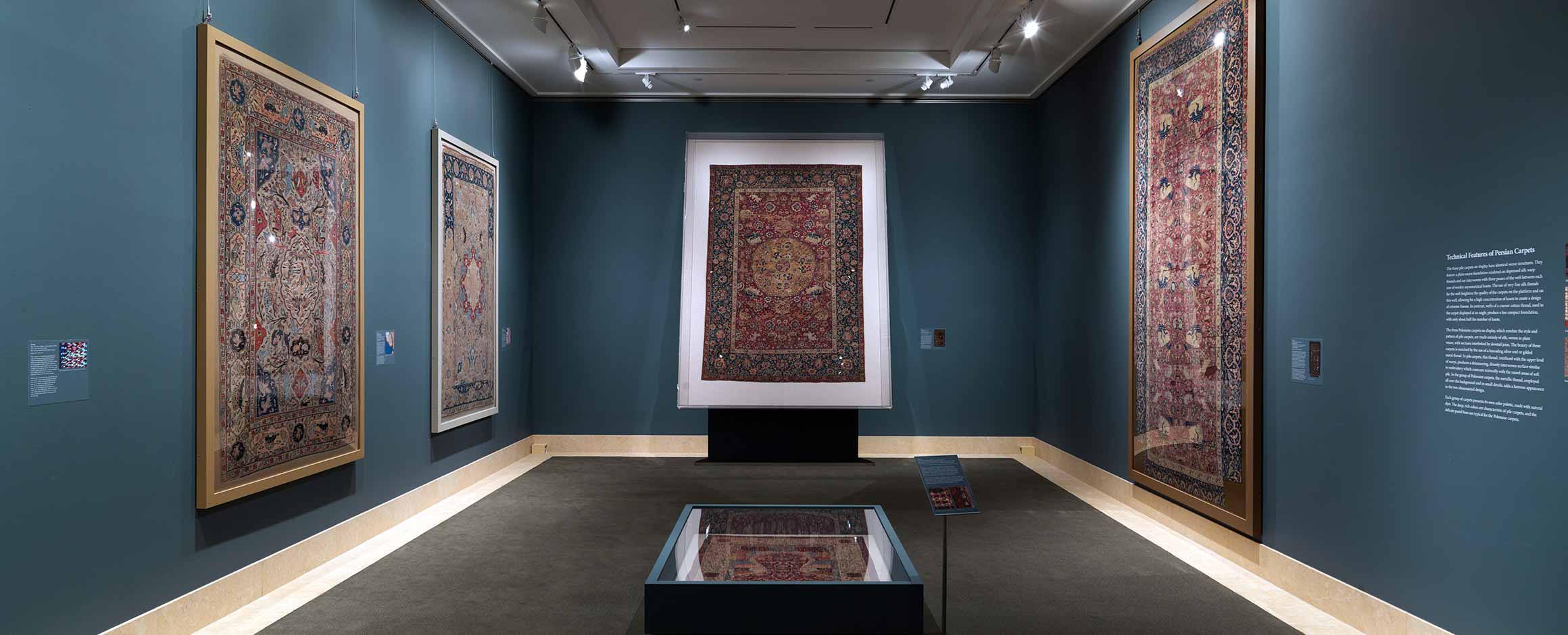 Installation view of "Carpets for Kings: Six Masterpieces of Iranian Weaving" with four large-scale carpets hung on dark teal walls in a small room.