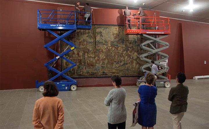 Art installers on electric lifts hanging a large-scale Renaissance tapestry with four people observing
