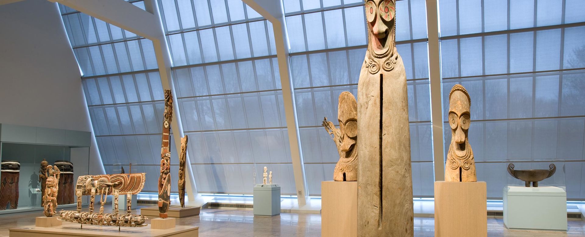 A large open gallery with a wall of windows; in the foreground is one very tall and two smaller wooden sculptures from Oceania