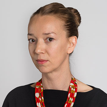 A woman with light brown hair pulled back in a bun, wearing a black, boat neck top and red and white beaded necklace