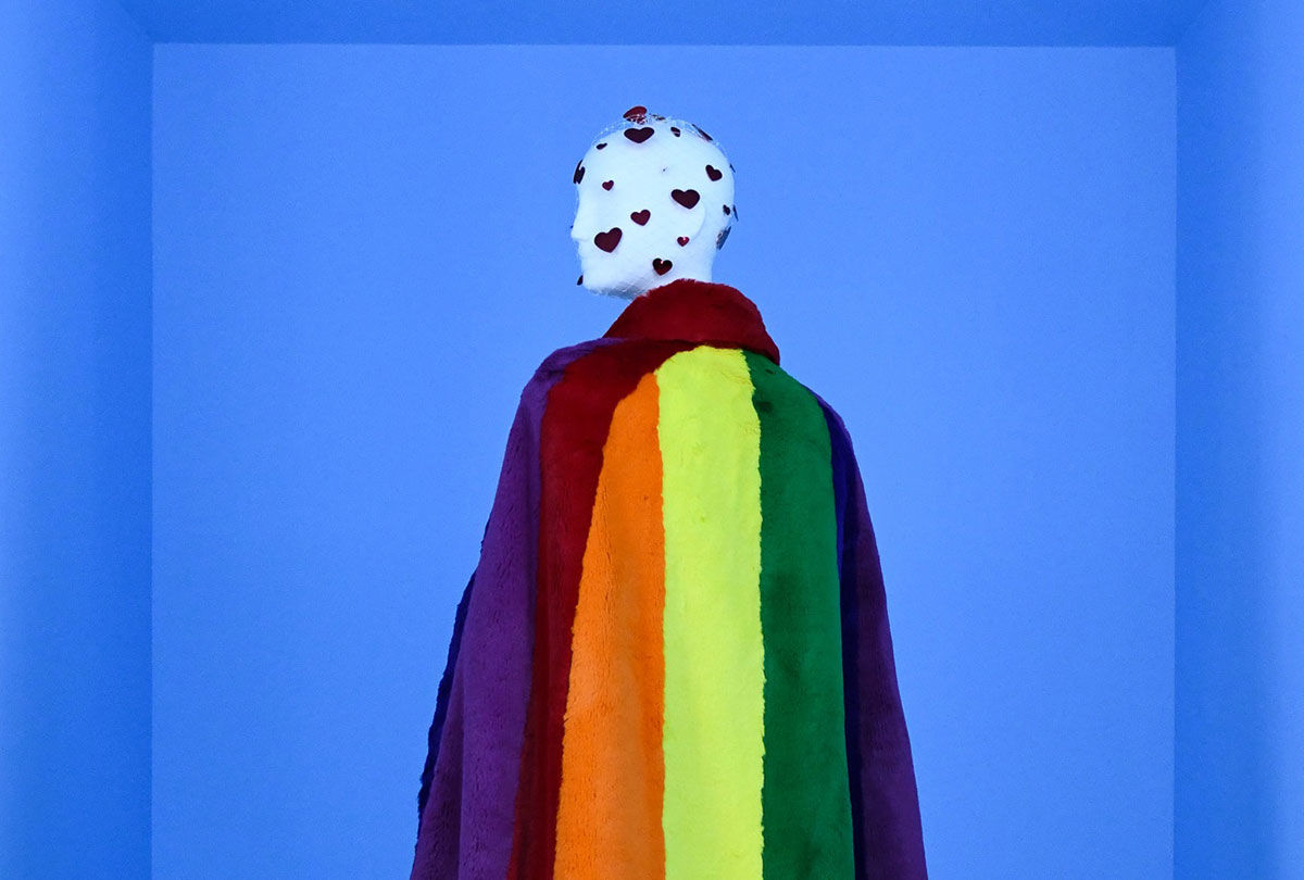 An installation view of a mannequin wearing a fuzzy Burberry cape in a bright rainbow design.