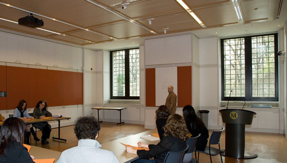 A well-lit modern, all-purpose room with white and brown panelling; a man is at the front of the room lecturing to a group at long tables set up in a U-shape