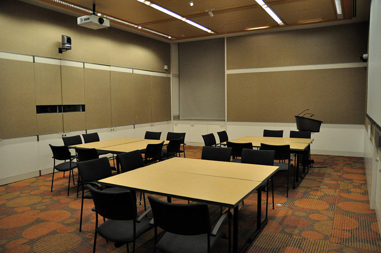 A small, comfortable, modern, carpeted room with beige walls and white paneling; the room is set with three large, square tables and chairs