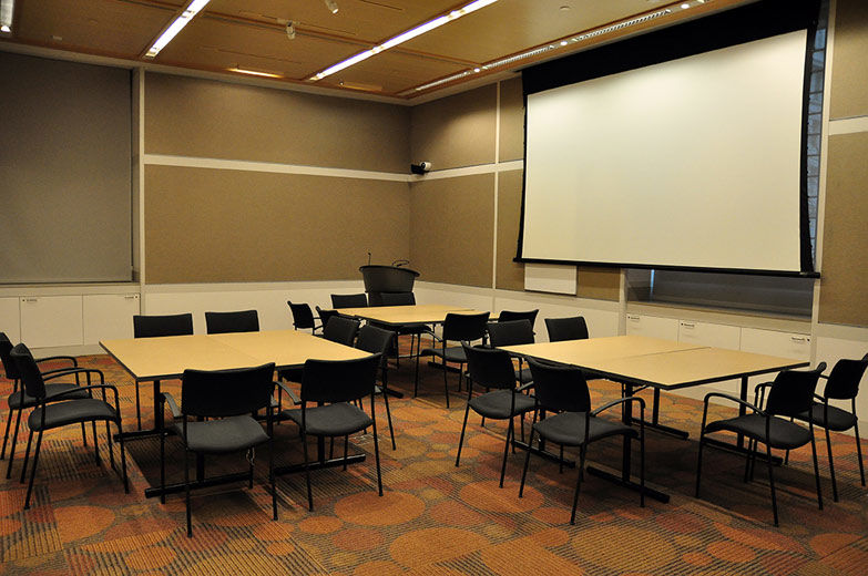 A small, comfortable, modern, carpeted room with beige walls and white paneling; the room is set with three large, square tables and chairs and the projection screen at the front of the room is lowered
