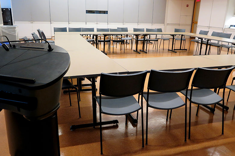 A large, comfortable, modern, all-purpose room with white walls covered in grey fabric paneling; the room is set with chairs and long tables in a square-shape; in the foreground is a lectern
