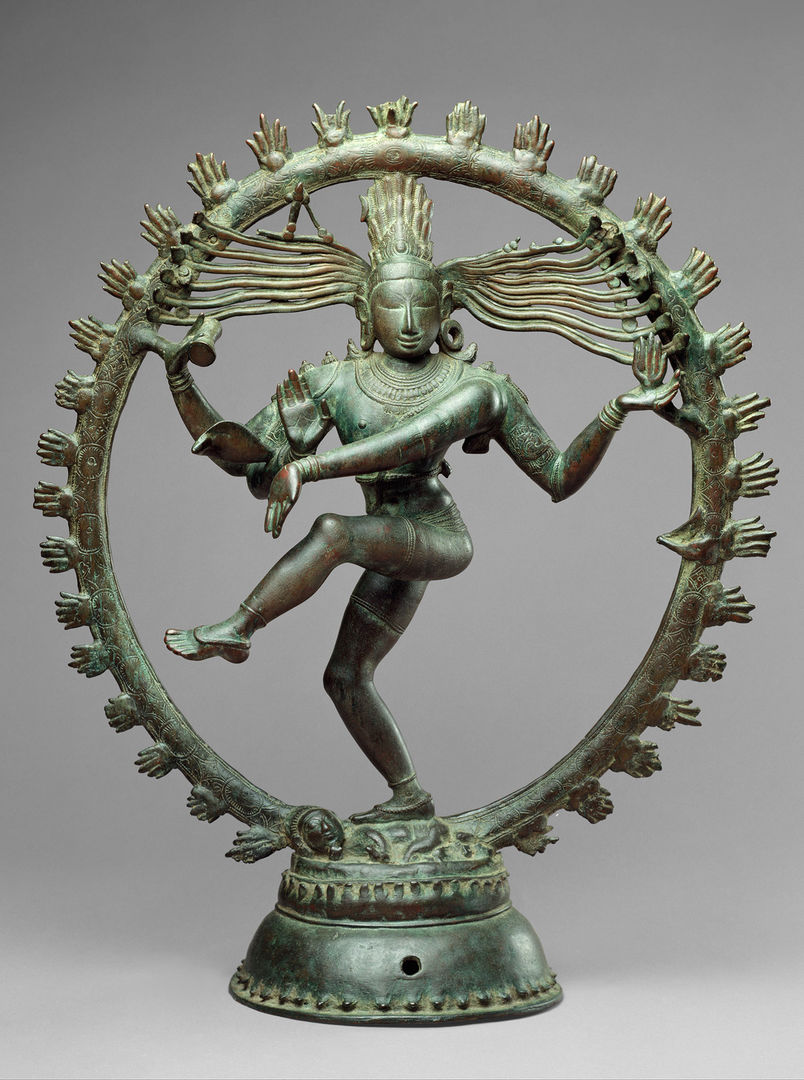 An oxidized copper sculpture of an Indian deity with four arms, standing on one leg dancing, encircled by a ring of stylized fire
