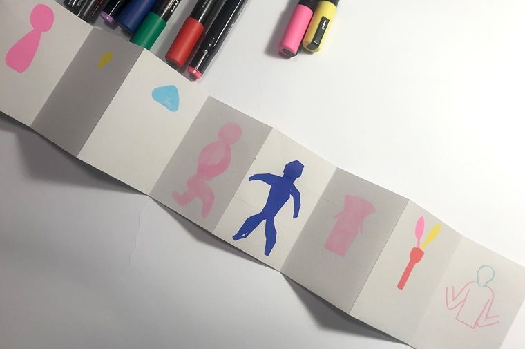 A photo of colored pencils and markers beside the paper strip, on which the the first layers of colored shapes have been drawn