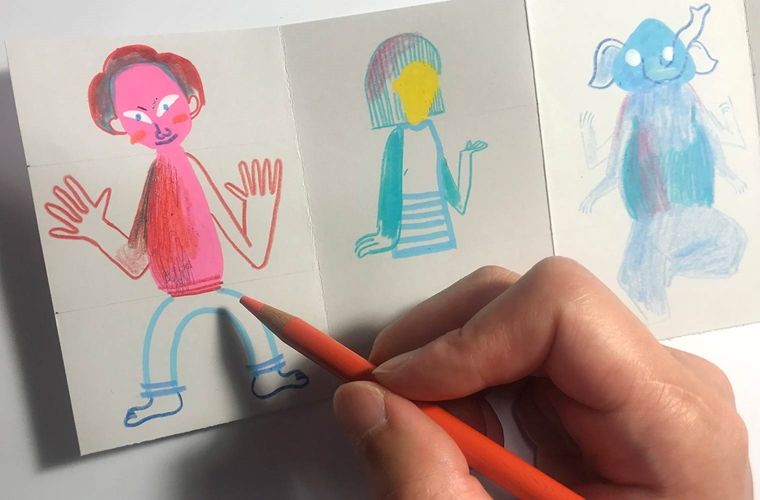 A hand holds an orange colored pencil and fills in one of the characters bodies with color on the paper.