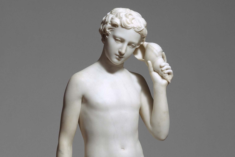 A white marvel carving of a muscular shirtless boy with curly hair holding a conch shell up to his ear and listening.