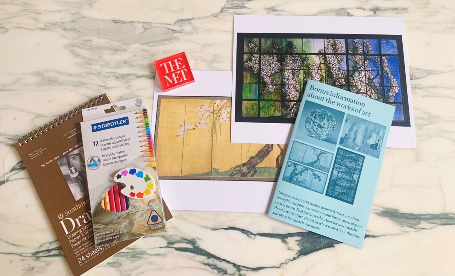 Art supplies, like colored pencils and an eraser, and printed images of art featuring flowering trees in nature spread out across a marble countertop.