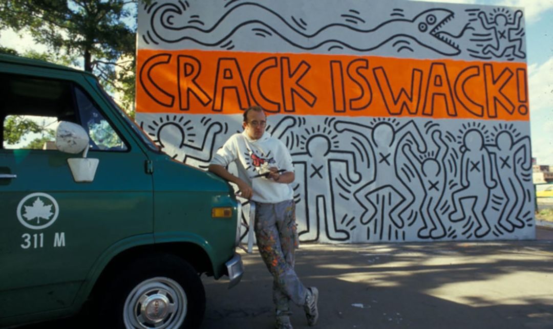 Archival photograph of the artist Keith Haring leaning on a New York City Department of Parks and Recreation truck with his mural called “Crack is Wack” appearing in the background; the mural features the title in big black outlined block letters against a neon orange background surrounded by illustrations of abstracted line figures moving expressively with one figure running away from a large snake.