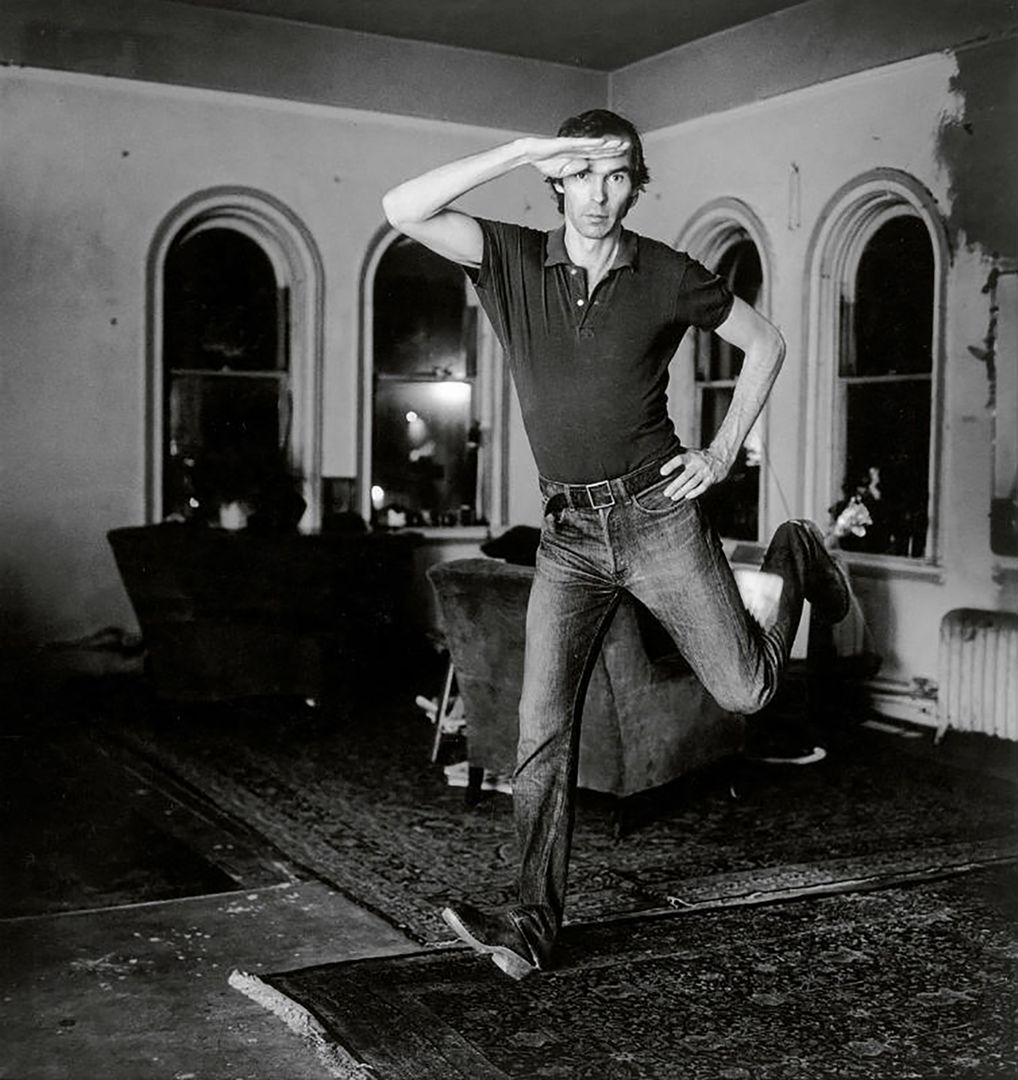 A black and white photograph of the photographer Peter Hujar jumping in a living room space at nighttime, decorated with Turkish rugs and arm chairs set against a corner bank of windows.
