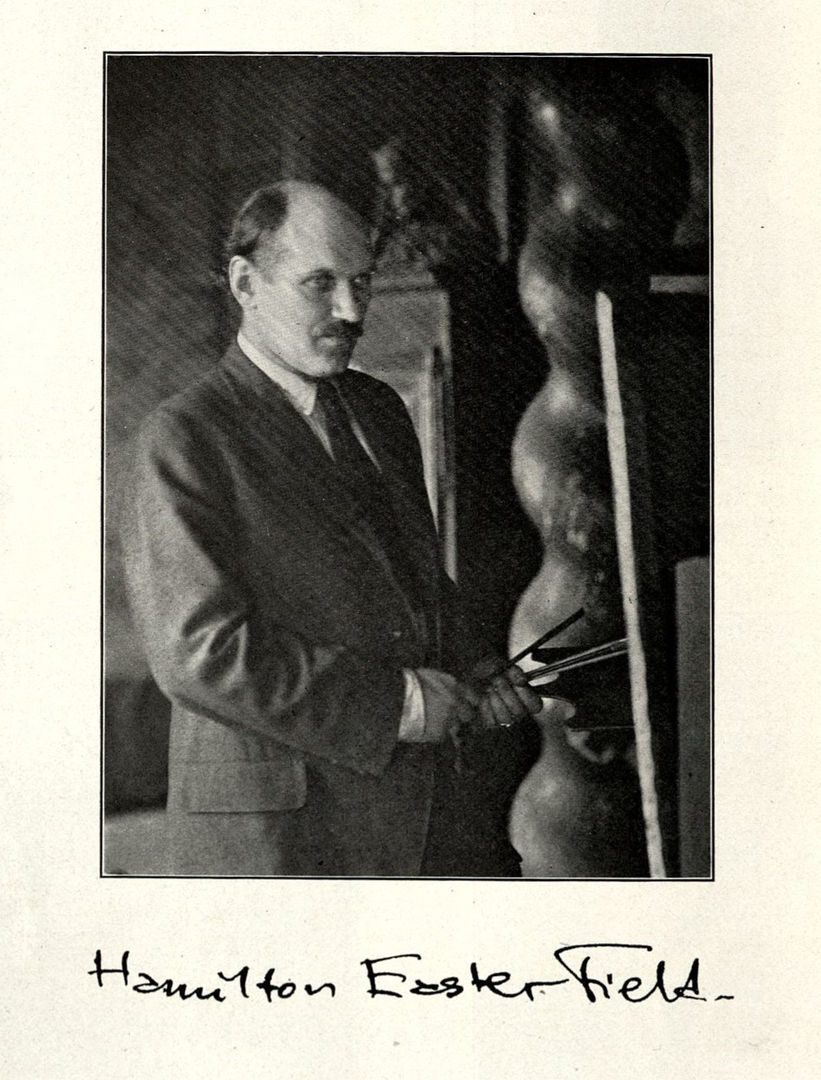 Black and white photograph of a white man in a suit with dark hair and a mustache. He his holding a bunch of paint brushes in front of a canvas. Below, there is a signature that says Hamilton Easter Field. 