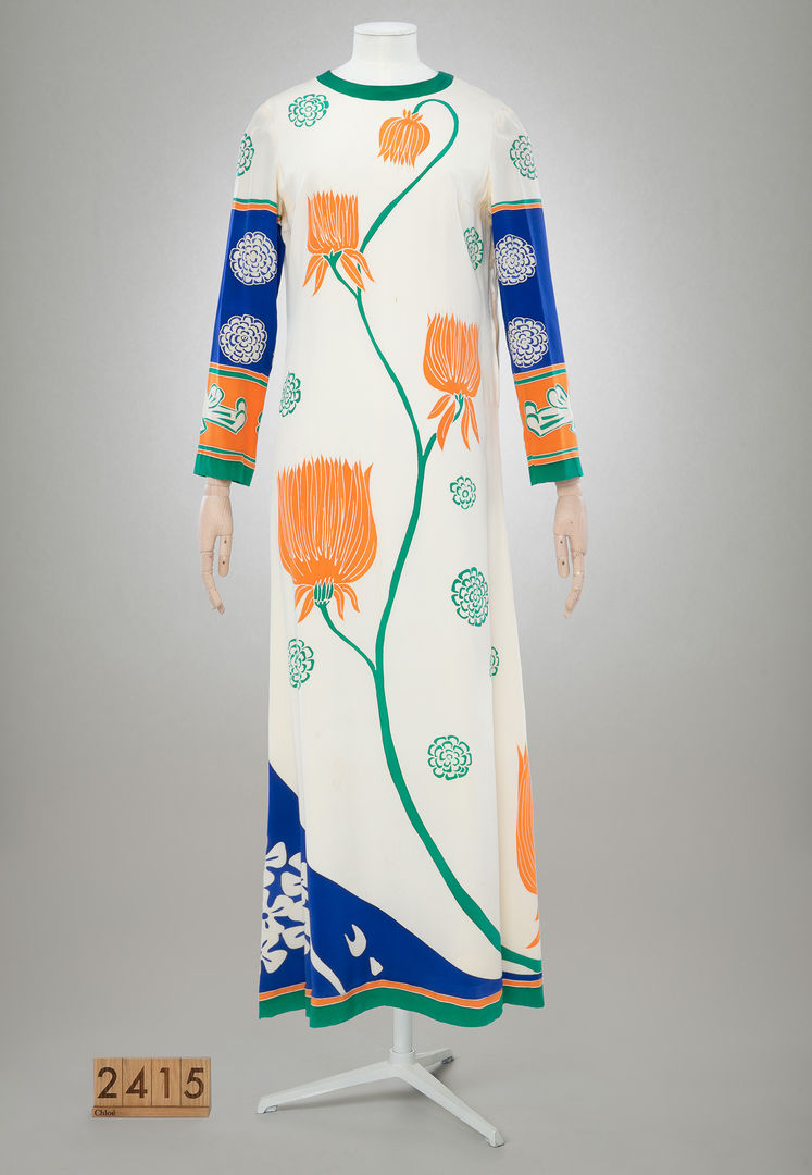 Image of a ankle length dress against a grey background. The dress features a very graphic design of four golden yellow flowers on green stems against a white background going up the body of the dress. The sleeves have blue and white patterns. The dress is fitted on a headless mannequin. 