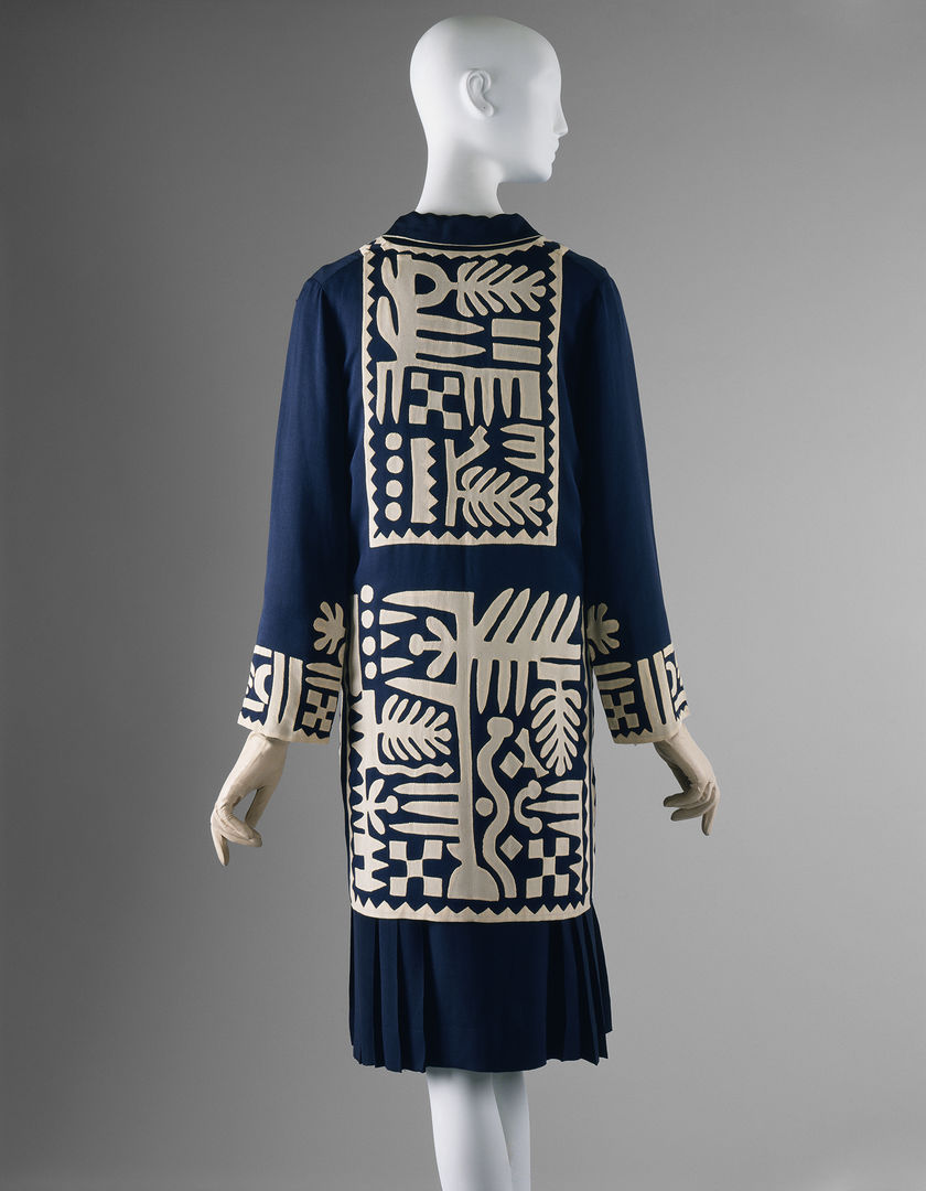 The back of a navy blue dress with beautiful white patterning on the back and lower part of the dress and around the wrists. The dress is against a grey background and fitted onto a mannequin with a head and hands. 