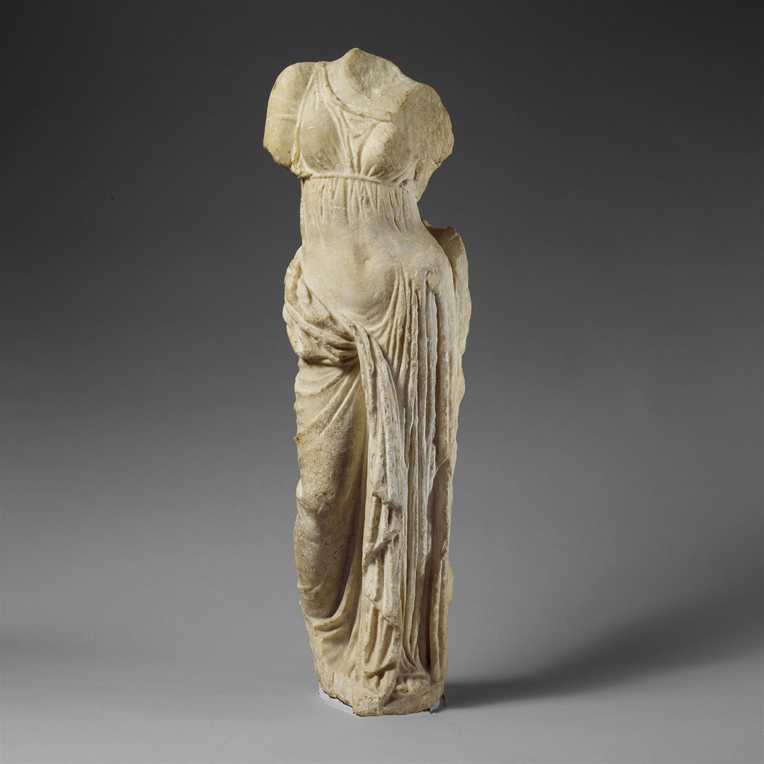 Old, ancient off white with dark stains marble sculpture of topless women with drapery covering her waist. The arms and head are missing and the statue sits against a grey gradient background with and is almost life-sized. 