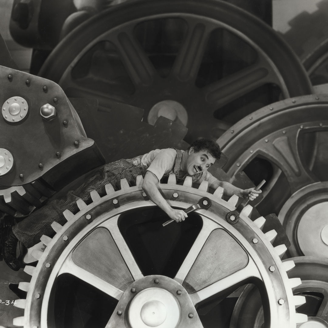 Black and white image of the actor Charlie Chaplin dressed as the tramp, a white man in overalls in this scenario and a mustache where he is trapped between massive factory cogs that he is riding while also trying to tighten the bolts with both of his hands at the same time. 