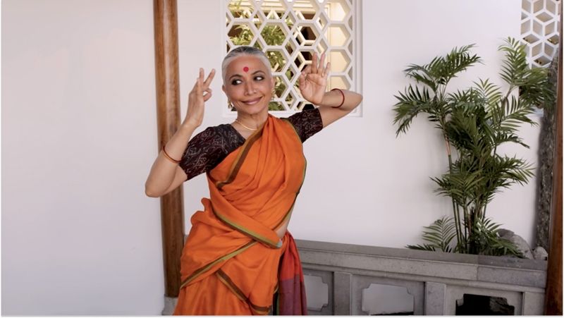 A woman stands wearing an orange sari and red bindi between her eyes. Her arms are held up near her head with her hands making a gesture. Behind her is a whitewall and green plant.