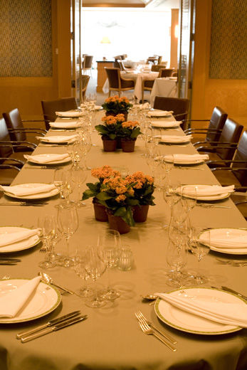 A casual-elegant private dining room with blond wood paneling and fabric covered walls, leather and chrome chairs; the table is set for fourteen with a casual service and simple potted-flower arrangements