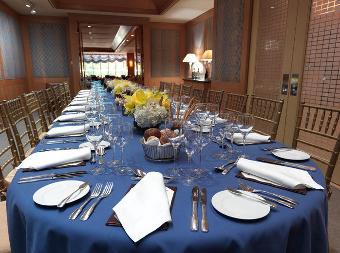 A casual-elegant private dining room with blond wood paneling and fabric covered walls, and padded fruitwood chairs; the table is set for eighteen with a casual service and low, elegant flower arrangements