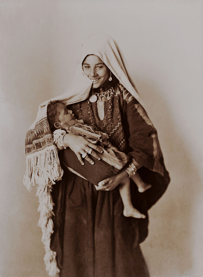 Black-and-white photograph of a smiling woman holding an infant in her arms. She wears a dark thobe with embroidery on the chest panel. A light-colored scarf is draped over her head, and she holds an edge of the scarf between her teeth.