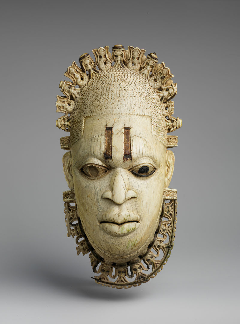 An ivory mask. Delicate carvings accent parts of the forehead and chin. A tiara and collar are decorated with carved mudfish and bearded Portuguese men.