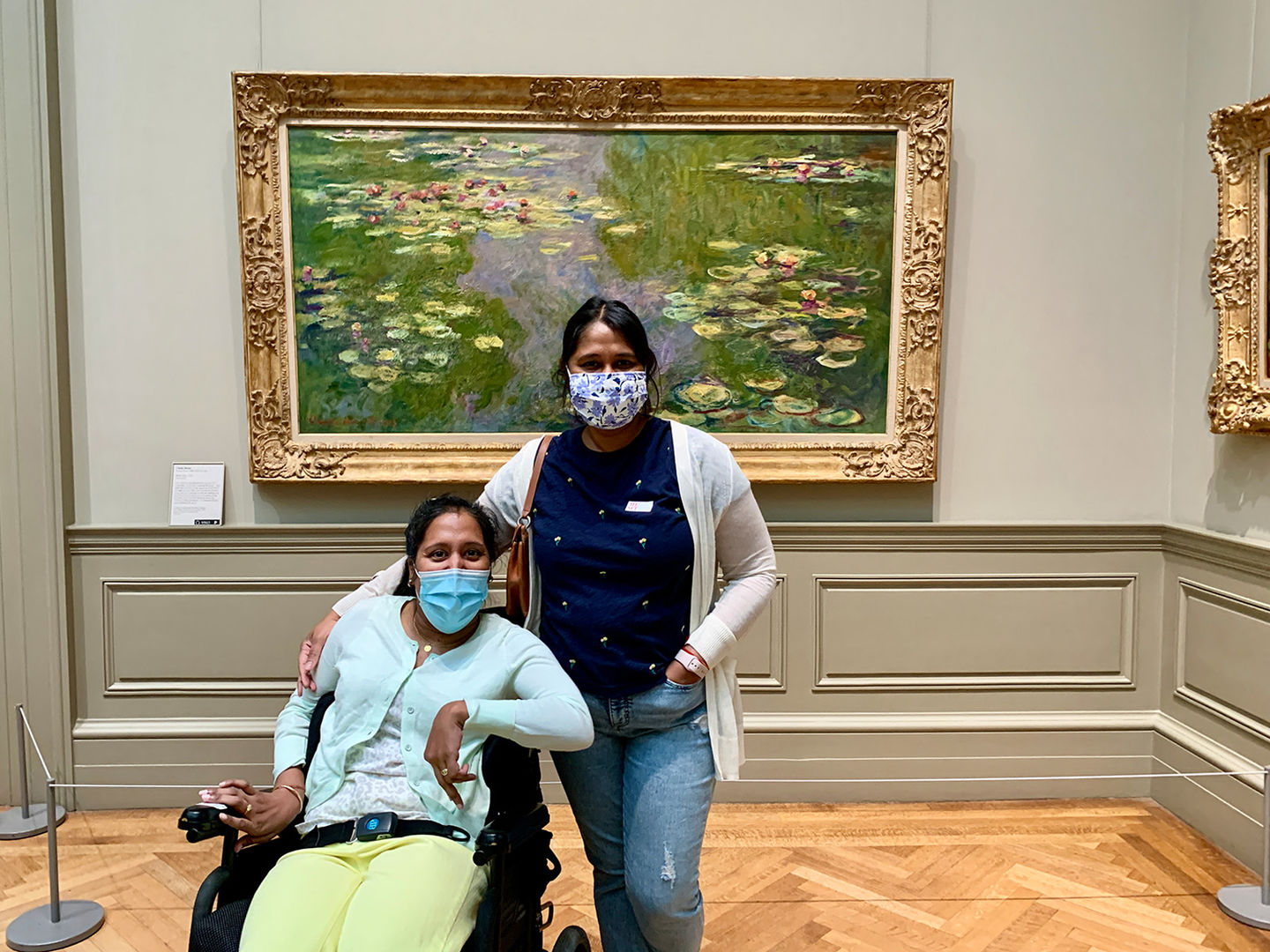 Portrait of two women, Annie and Lakshmee, in the European Painting galleries at The Met 5th Avenue. The women are medium-dark skinned and one of them is a wheelchair user. 