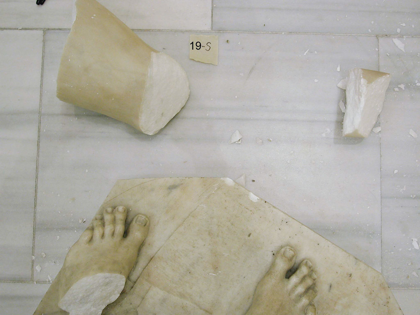 Fragments of Adam against a gray tile floor. Larger pieces show the breakage at his feet and parts of his appendages, and are surrounded by tiny pieces of marble.