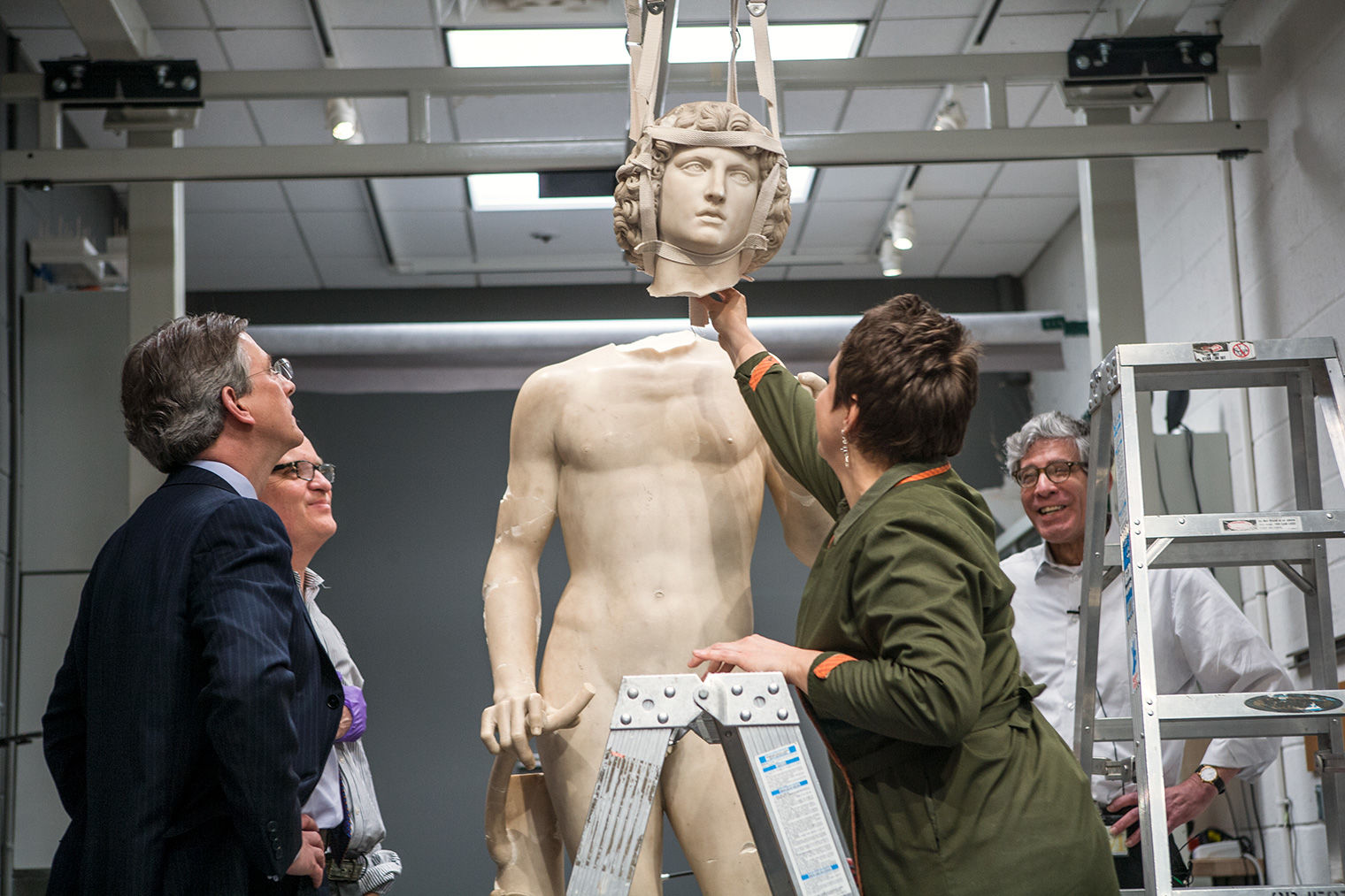 In a gray-walled room, four people stand around Adam as his head, hanging from a harness, is reattached to his body.