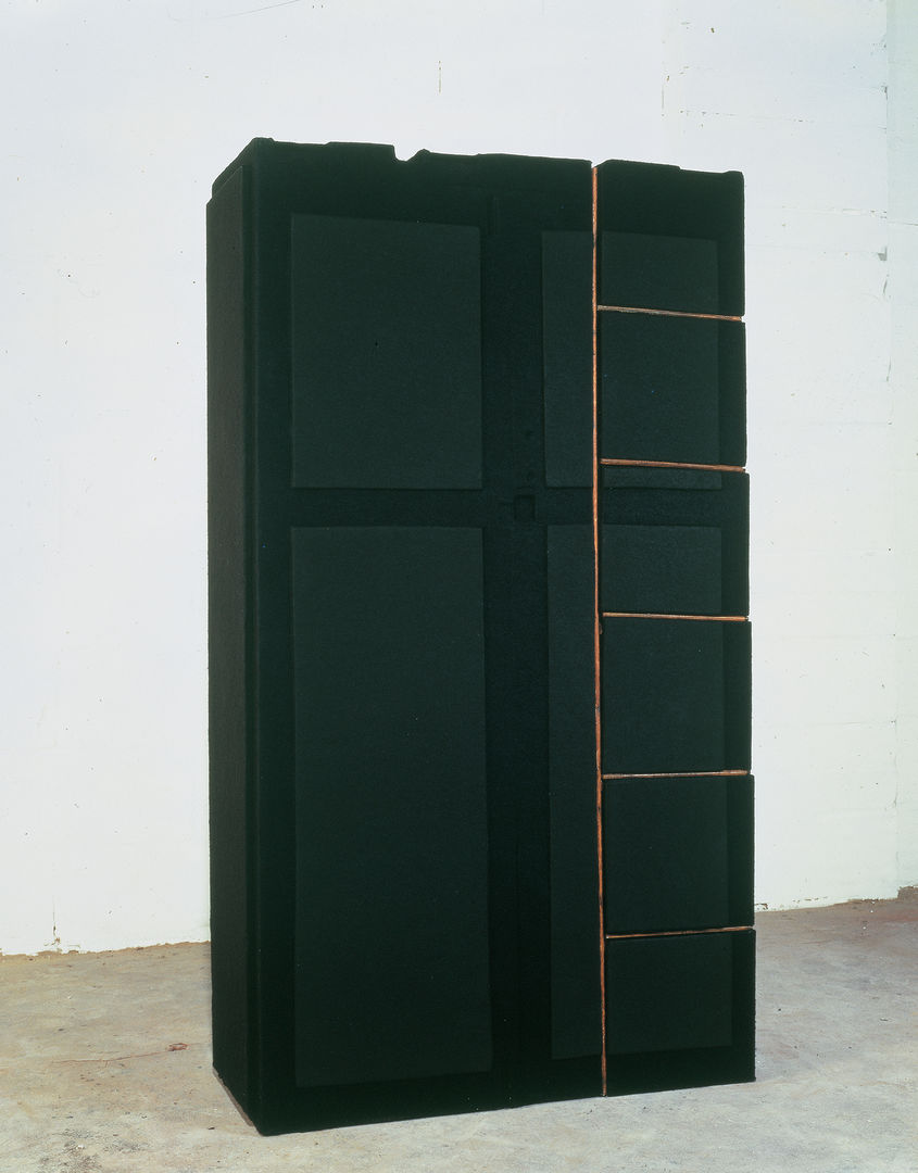 Large black wardrobe, set on a concrete floor against a white wall. Dark gray paneling and orange grooves accent the doors of the wardrobe.