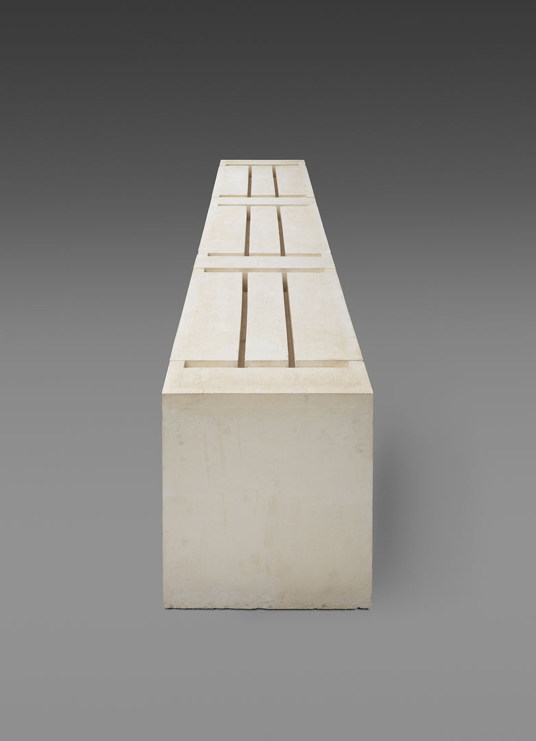 Bulky, rectangular plaster sculpture against a gray background. Grooves on the upper side of the sculpture outline the underside of three tables. 