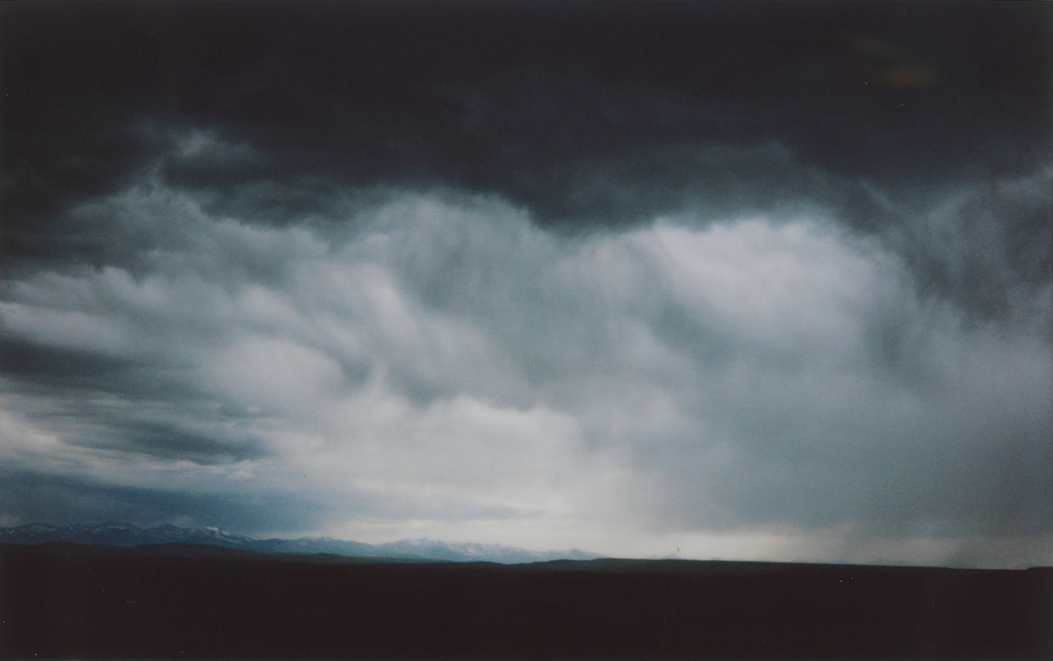 Landscape photograph. Dark blue, stormy clouds give way to hazier gray clouds. In the distance, snow-capped, blue mountains. The ground throughout the shot is dark gray.