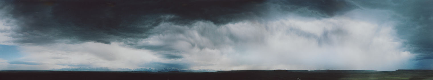 Long landscape photograph showing dark blue, stormy clouds gradually giving way to white and gray clouds in the distance, with patches of blue skies peeking through. The ground is darkened by the clouds, but the snow-capped mountains in the distance are cast in blue light.