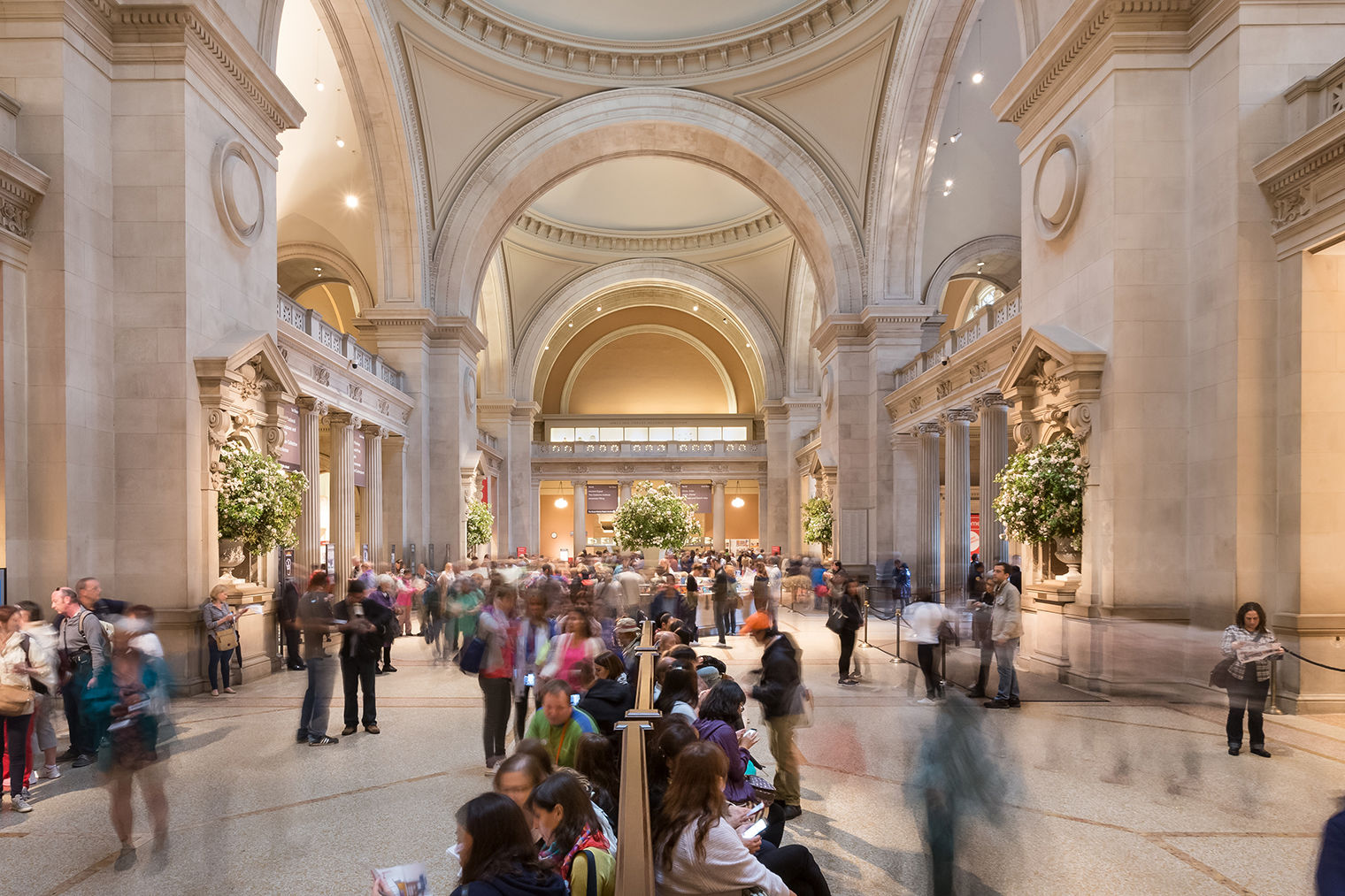 A bustling photo: people are gathered at the information desk, looking at maps, and resting in seats, against the majestic columns and domes of The Met's Great Hall.