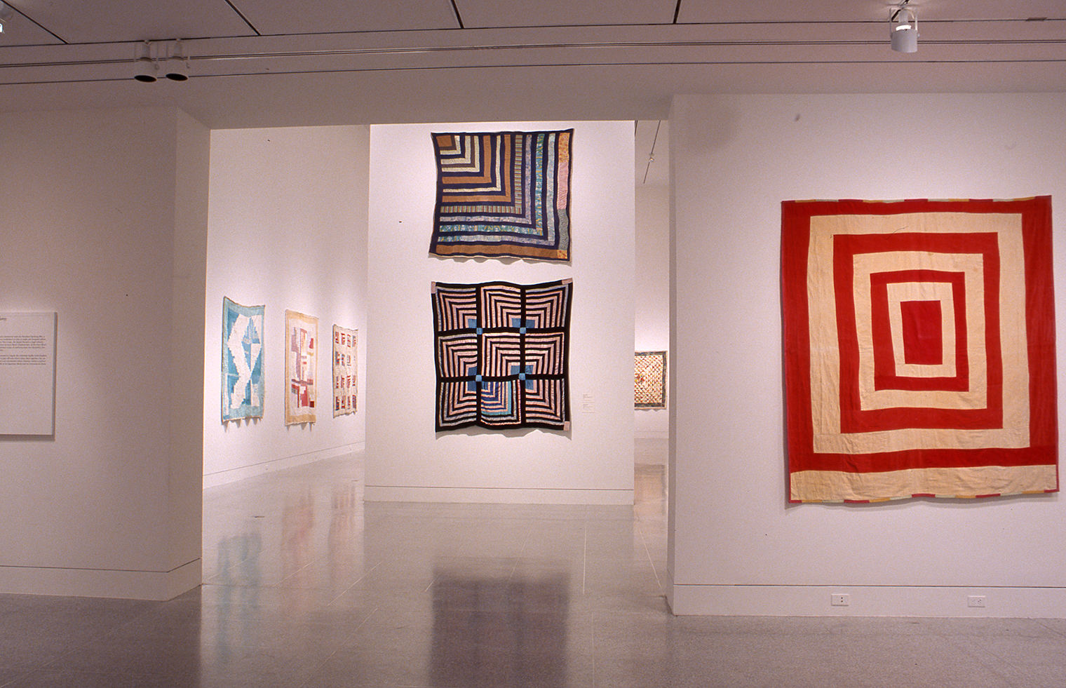 Installation shot of Gee's Bend exhibition at the MFA Houston in 2002. At right, and through a doorway in the background, multicolored quilts hang on white walls.