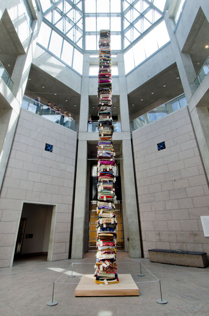 In a high-ceilinged, brightly-lit atrium, an installation of stacked, colorful blankets stretches multiple stories in height. A small group of people are viewing the installation from an upper floor.